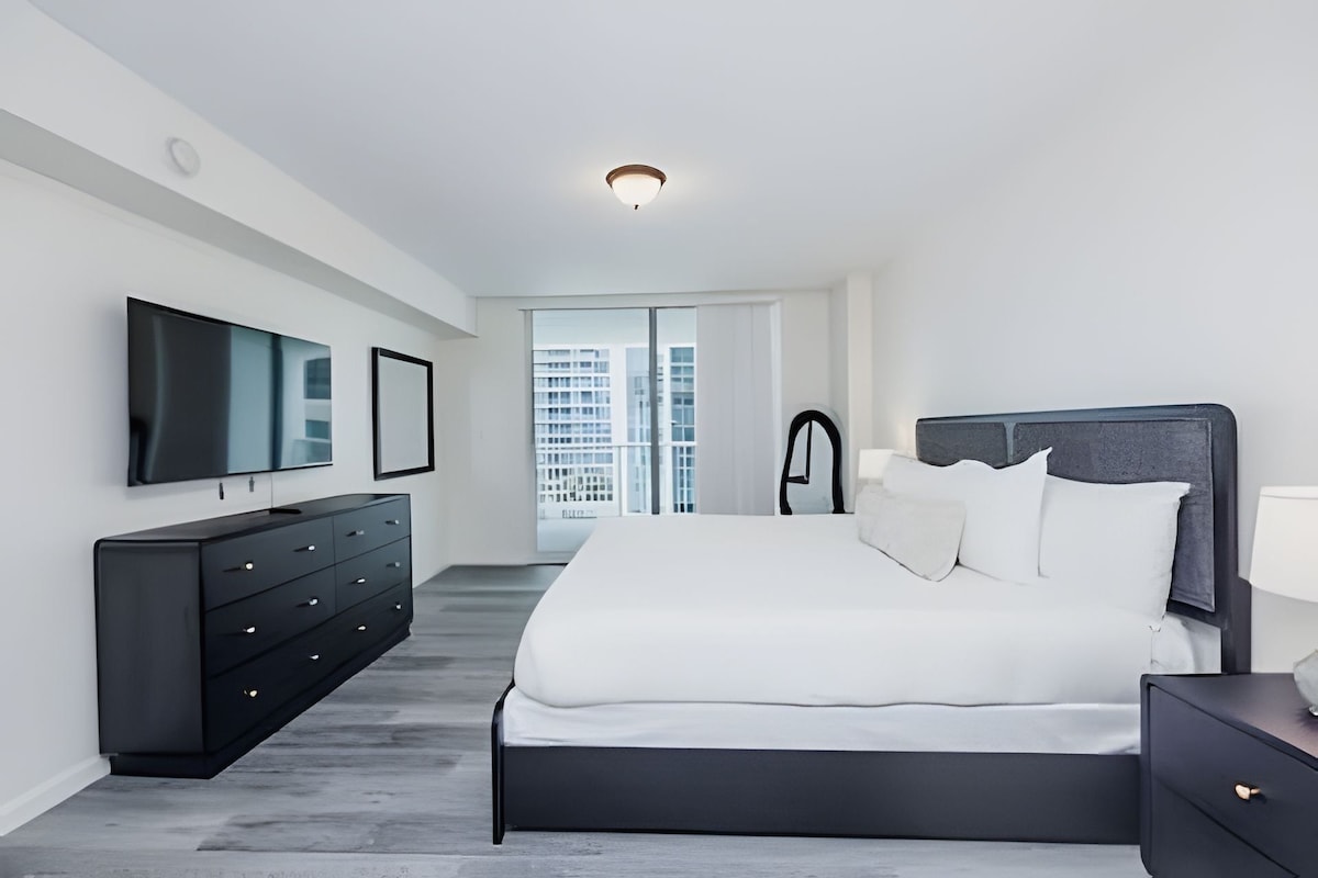 Quarto moderno com uma cama grande, televisão de tela plana, cômoda e vista para a cidade pela janela.