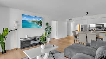 A modern living room with a flat-screen TV, a sofa, a coffee table, and a kitchen in the background.