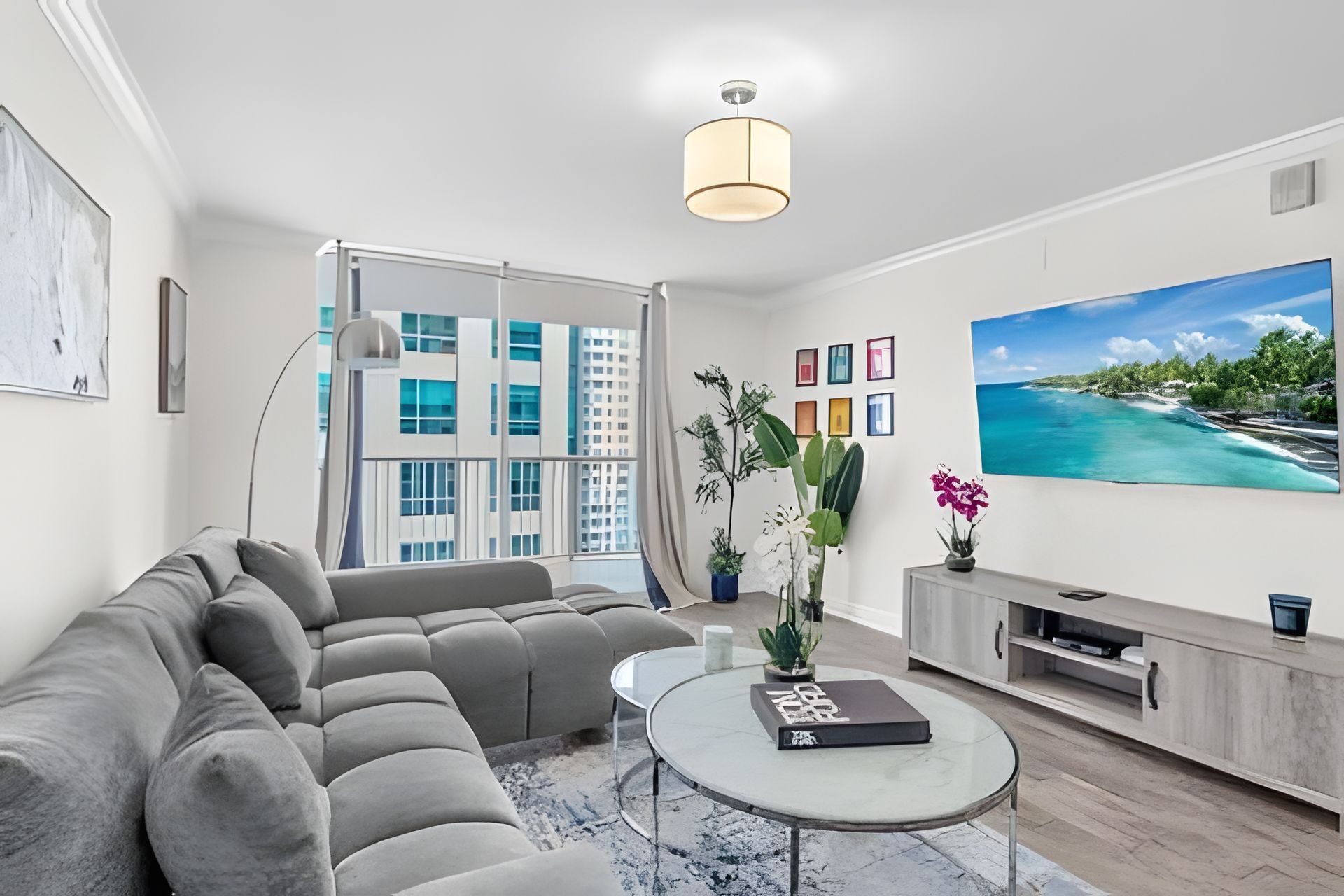 A modern living room with a sectional sofa, a glass coffee table, a flat-screen TV, and a large window with a view of buildings.
