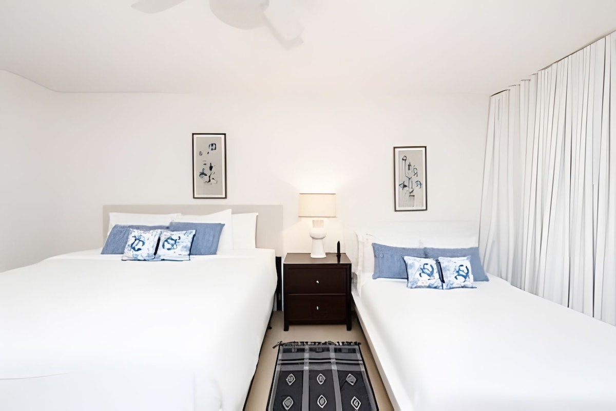Quarto de hotel com duas camas, uma mesa de cabeceira, uma luminária e obras de arte nas paredes.