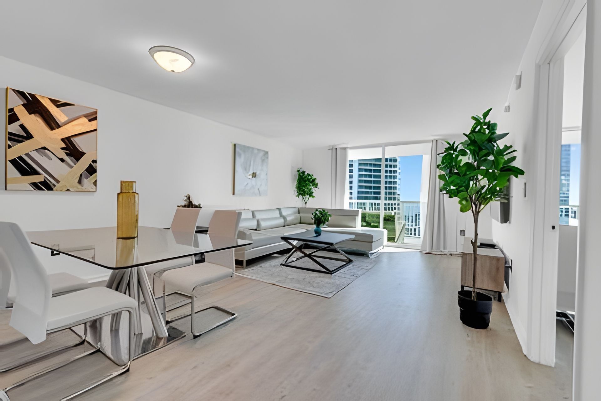 A modern living room with a glass dining table, white sofas, and a large window offering a city view.