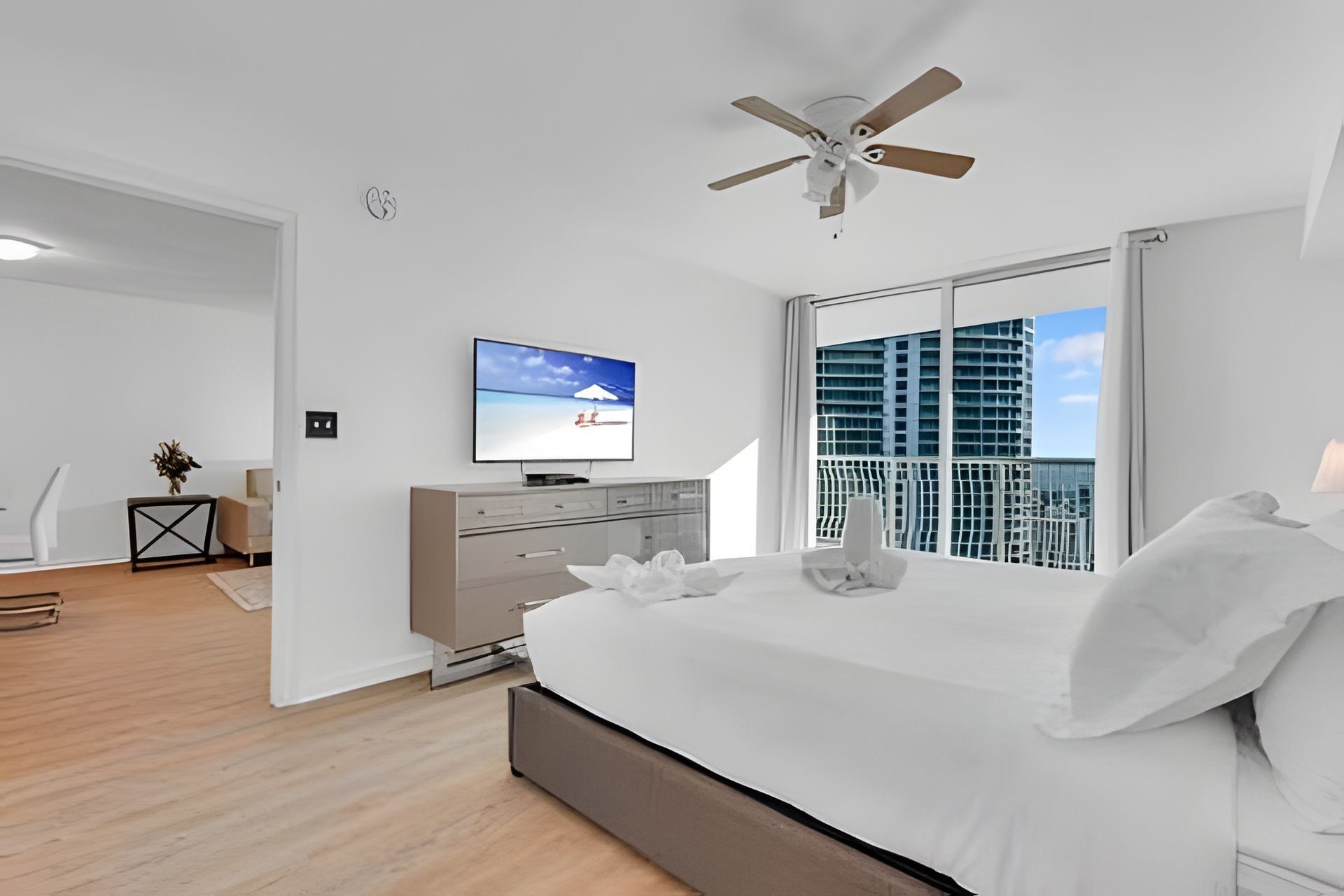 Quarto moderno com cama, televisão, ventoinha no teto e vista da cidade através de amplas janelas.