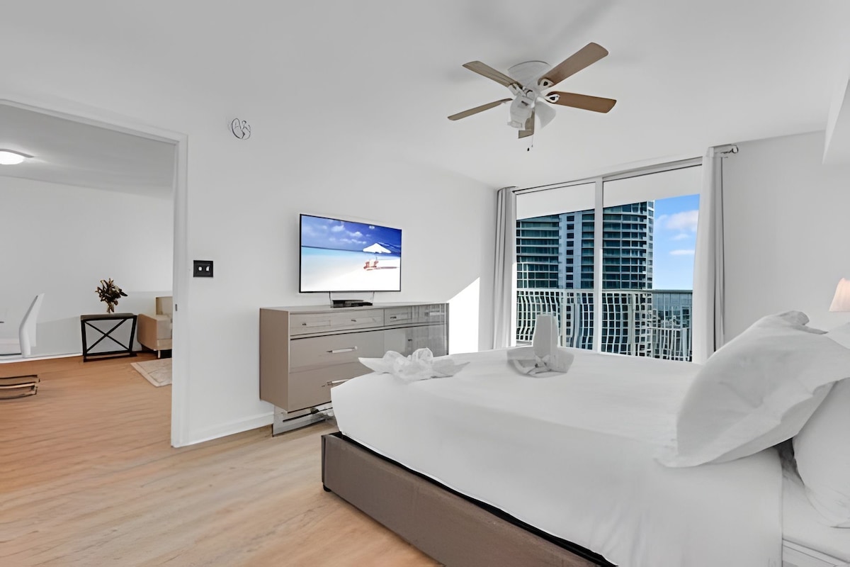 Quarto moderno com cama, televisão, ventoinha no teto e vista da cidade através de amplas janelas.