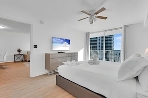 Quarto moderno com cama, televisão, ventoinha no teto e vista da cidade através de amplas janelas.