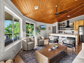 Un'ampia zona giorno con soffitto in legno, ampie finestre, una cucina dotata di elettrodomestici moderni e un'area pranzo con arredi contemporanei.