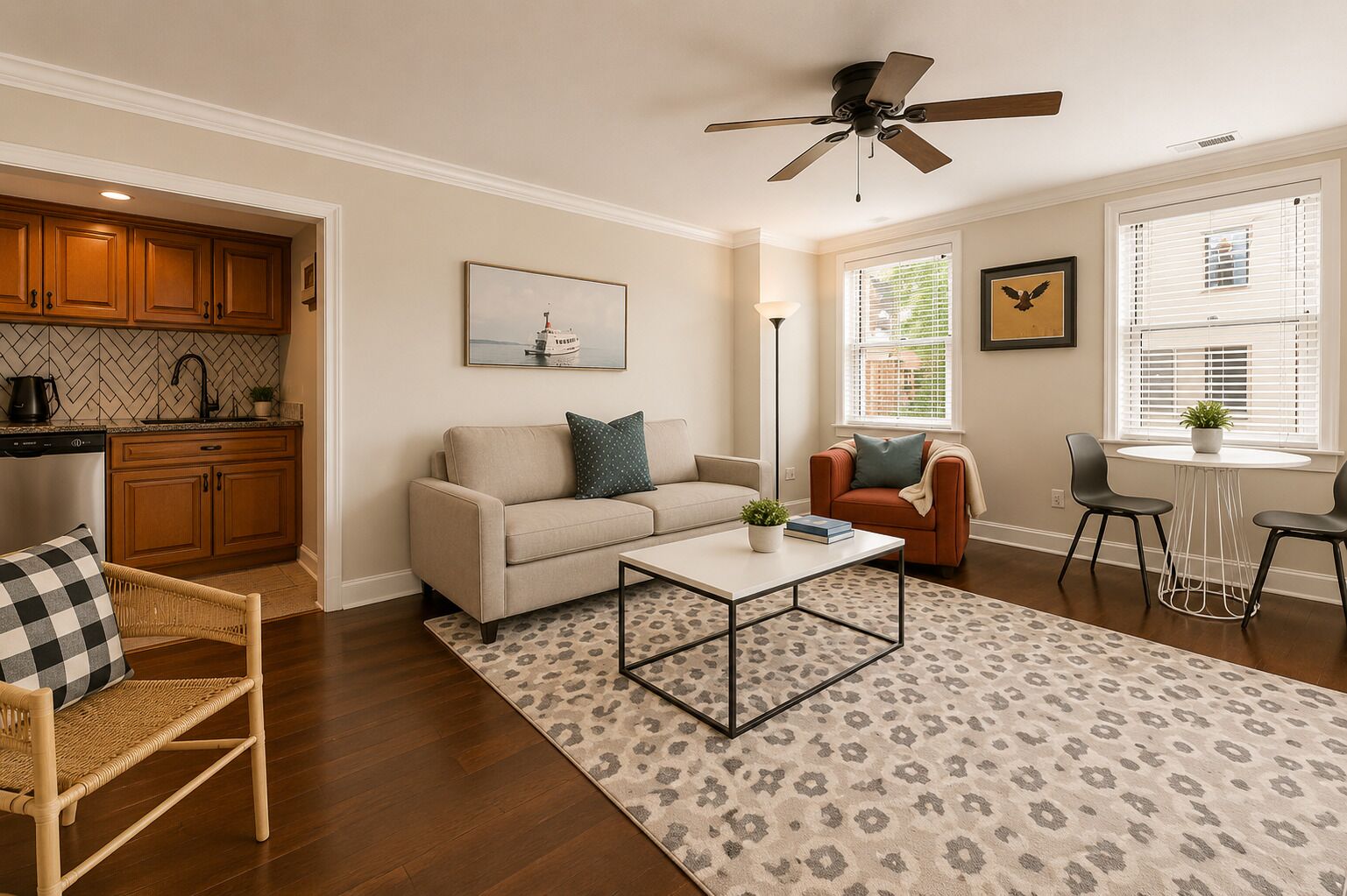 A living room with a sofa, coffee table, and chairs, a kitchen with wooden cabinets, and a ceiling fan.