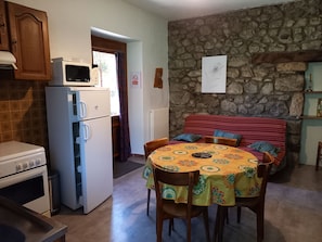Eine Küche mit Kühlschrank, Herd und Mikrowelle. Ein Esstisch mit Stühlen und ein rot gestreiftes Sofa sind ebenfalls vorhanden. Im Hintergrund sind eine Steinwand und eine Tür zu sehen.