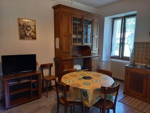 Ein Essbereich mit einem runden Tisch, Stühlen, einem Fernseher auf einem Schrank, einer Vitrine und einem Fenster mit Vorhängen.