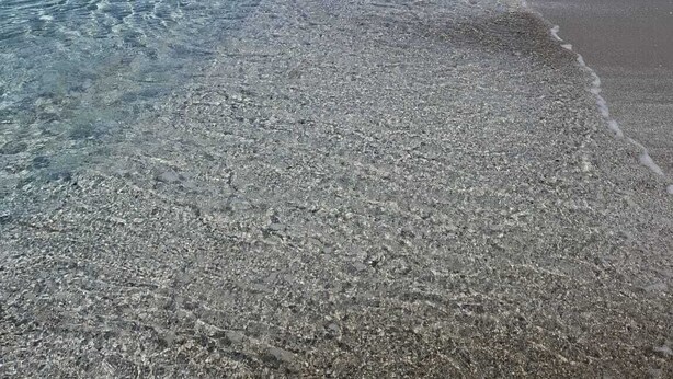 En strand med klart vand, en stenet kystlinje og klippeformationer i baggrunden.