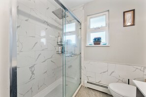 Un baño con cabina de ducha de vidrio, inodoro, ventana con vistas y paredes con diseño de mármol.