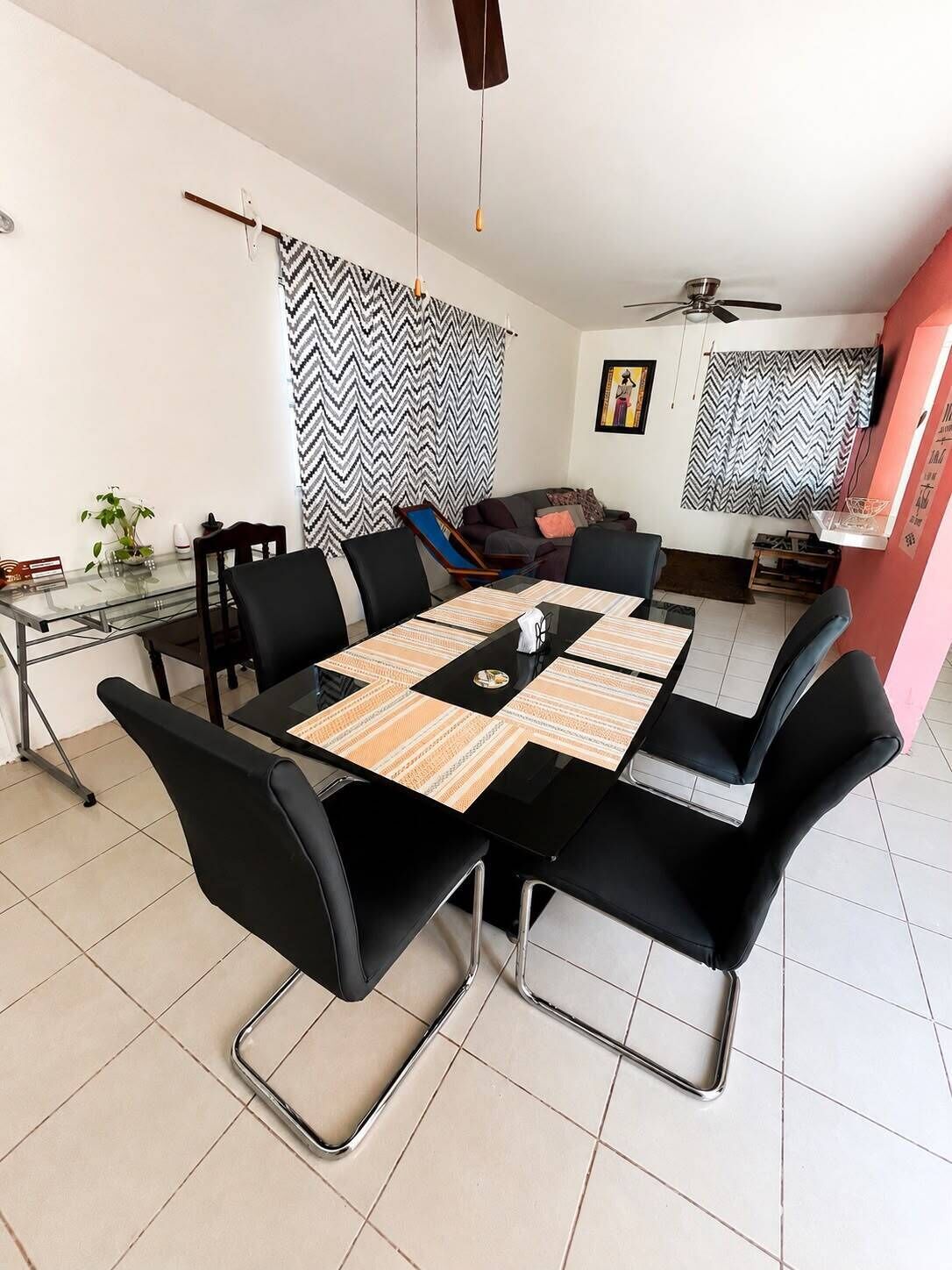 Un comedor con una mesa de vidrio, sillas negras, un ventilador de techo y un sofá al fondo.