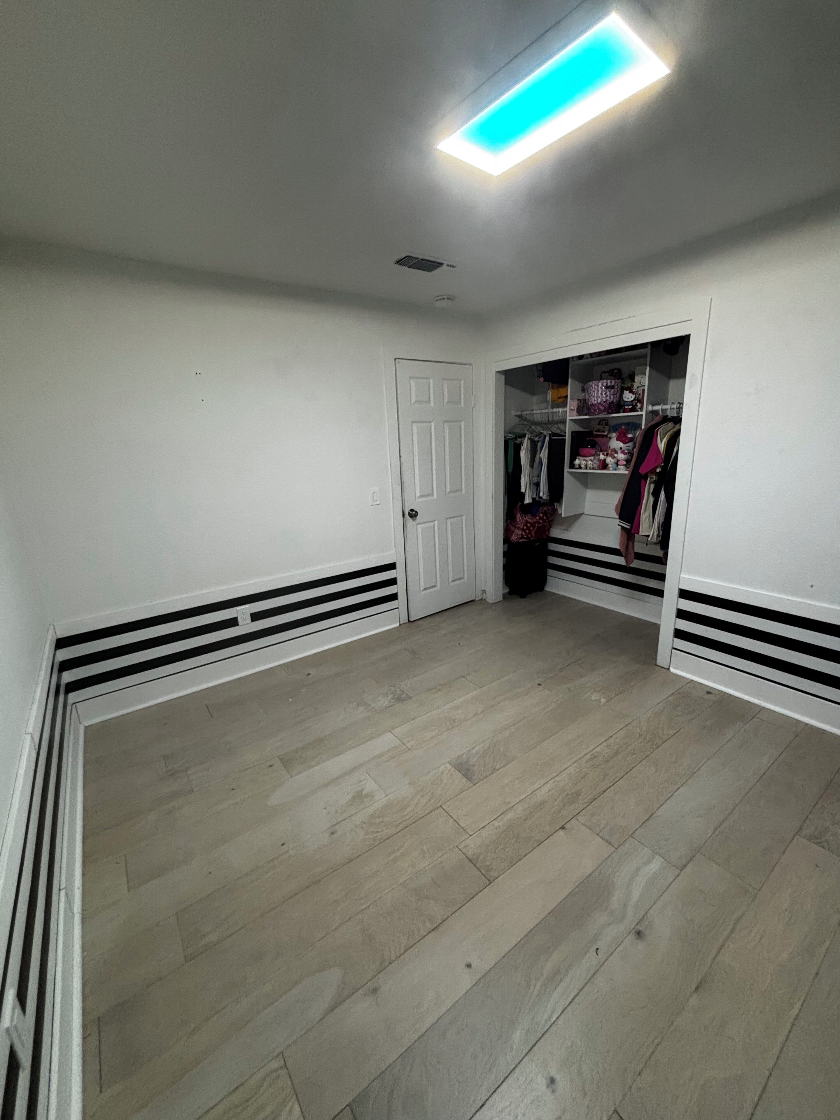 A room with a wooden floor, white walls, a closet with hanging clothes, and a door.