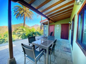 A covered patio with a table and chairs, a view of palm trees and mountains, and a door with a sign. - El pirguan, holiday house (Vallehermoso)