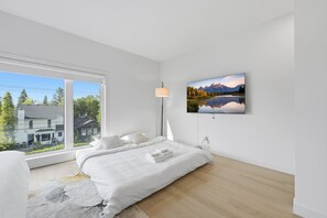 Un dormitorio moderno con un ventanal grande, una cama con ropa de cama blanca, un televisor de pantalla plana y una lámpara.