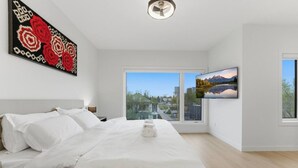 A modern bedroom with a large bed, a view of a cityscape through a window, and a painting on the wall. - 4 Ensuites | Private Rooftop | Bar Lounge | Slps 8 (Calgary)