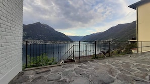 En balkong med stenbeläggning och ett metallräcke, med utsikt över en sjö och berg.