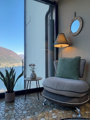 Quarto com vista para um lago e montanhas, uma cadeira redonda com almofada, uma mesa pequena com um vaso e um abajur.