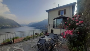 A terrace with a view of a lake and mountains, a building with a balcony, and a rose bush. - Villa top view Lake Como (Faggeto Lario)