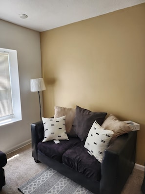 En stue med en mørklilla sofa, beige og sorte puder, en gulvlampe og en tæppe med mønster.