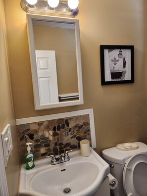 A bathroom with a white sink, a mirror, a framed picture, a toilet, and a wall-mounted light fixture.
