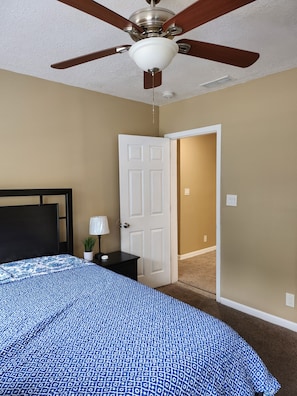 A bedroom with a bed, a nightstand, a lamp, a ceiling fan, and a door leading to another room.