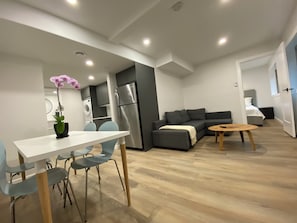 Ein moderner Wohnraum mit Essbereich, einer Couch, einem kleinen Tisch und einer Küchenzeile, inklusive Kühlschrank und Waschmaschine.