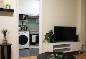 Uma cozinha moderna com micro-ondas, forno e máquina de lavar roupa, uma TV preta sobre um suporte branco e um pequeno vaso com planta.