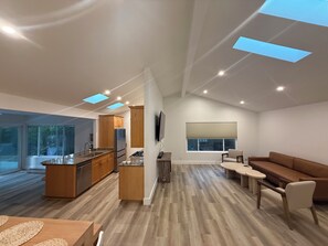 Eine moderne Küche mit Kochinsel, ein Essbereich mit Tisch und Stühlen, ein Wohnzimmer mit Sofa und ein großes Fenster mit Jalousien.