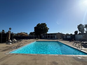 Una piscina rectangular con agua azul cristalina, rodeada de sillas de descanso y sombrillas, con un edificio al fondo.