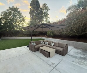 Eine Terrasse mit einer Eckcouch und einem Tisch unter einem großen Sonnenschirm, umgeben von Grünpflanzen und Bäumen.