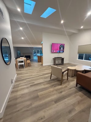 A spacious living area with a wooden floor, a TV mounted on the wall, and a large window.