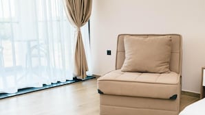 Un sofá color crema con un cojín en una habitación con piso de madera y un ventanal con cortinas translúcidas.