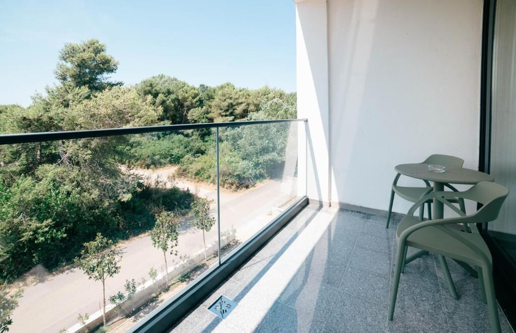A balcony with a glass railing, a table with chairs, and a view of trees and a road.
