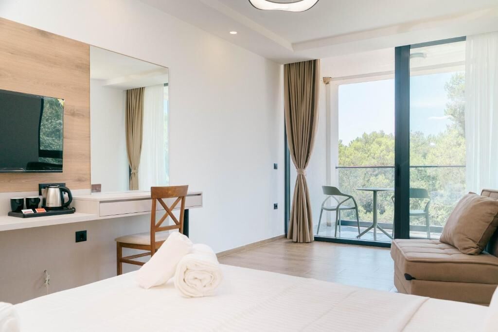 A hotel room with a bed, a desk with a coffee maker, a chair, a sofa, a balcony with a view, and a television mounted on the wall.