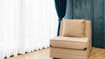 Un sofá beige individual con un cojín en una habitación con suelo de madera, cabecera acolchada azul y cortinas blancas.