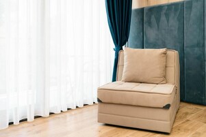 Un sofá beige individual con un cojín en una habitación con suelo de madera, cabecera acolchada azul y cortinas blancas.