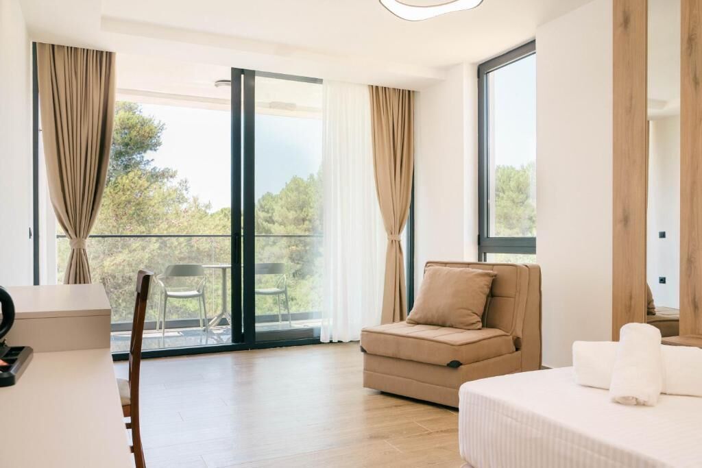 A modern hotel room with a sofa, a bed, a desk, and a view of the outdoors through large windows.