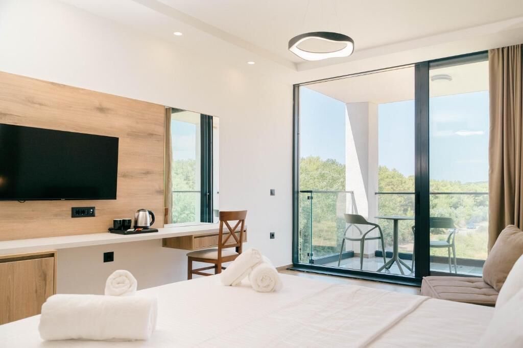 Een moderne hotelkamer met een flatscreen televisie, een bureau, een stoel, een bed met witte lakens en een balkon met een tafel en stoelen.