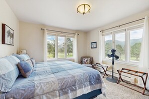 Ein Schlafzimmer mit einem großen Bett, einem Standventilator, einem Sessel und Blick ins Freie durch große Fenster.