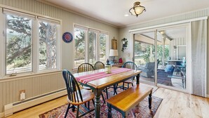 A dining area with a wooden table, chairs, and a bench. There are large windows and a view of trees outside. - Comfortable Cottage With Wifi, Washer-dryer - Golf & Mountain Views (Estes Park)