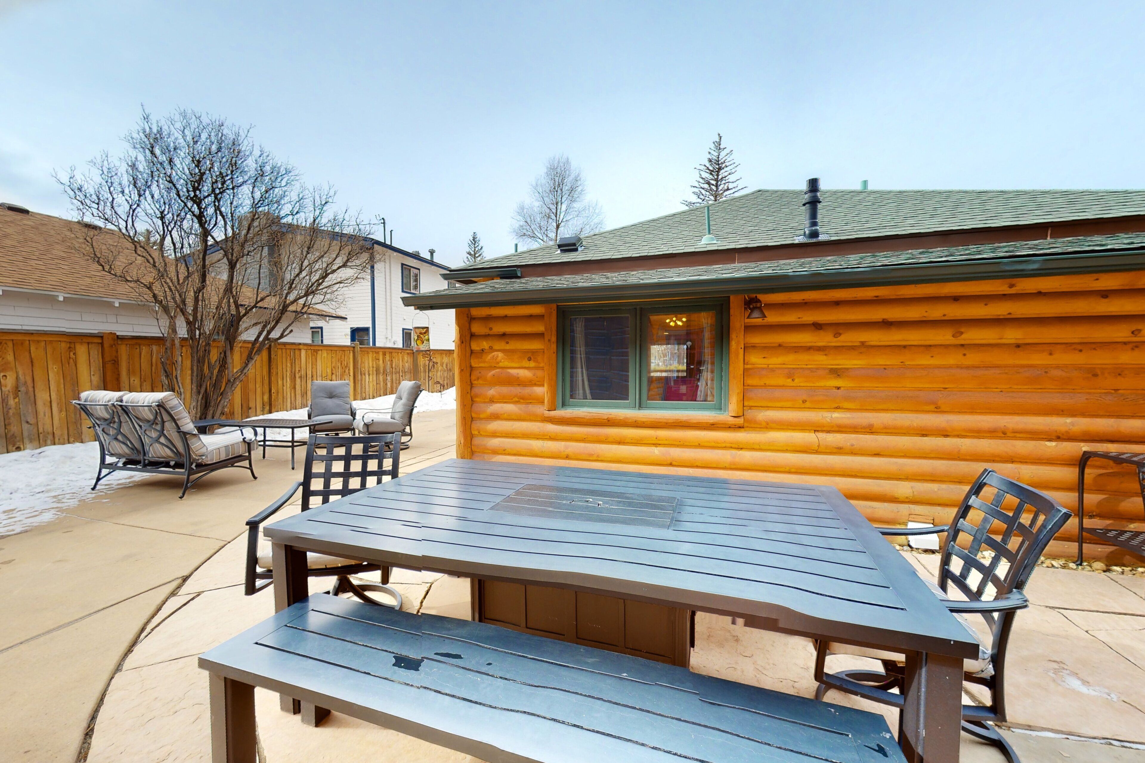 A log cabin with a patio area featuring a table and chairs, a wooden fence, and a view of the surrounding landscape.