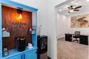 A coffee station with a coffee maker and water dispenser, a desk with a chair, and a ceiling fan in a room with a painting on the wall. - Remodeled, Dog-friendly Cabin in Heart of Downtown With Patio (Estes Park)