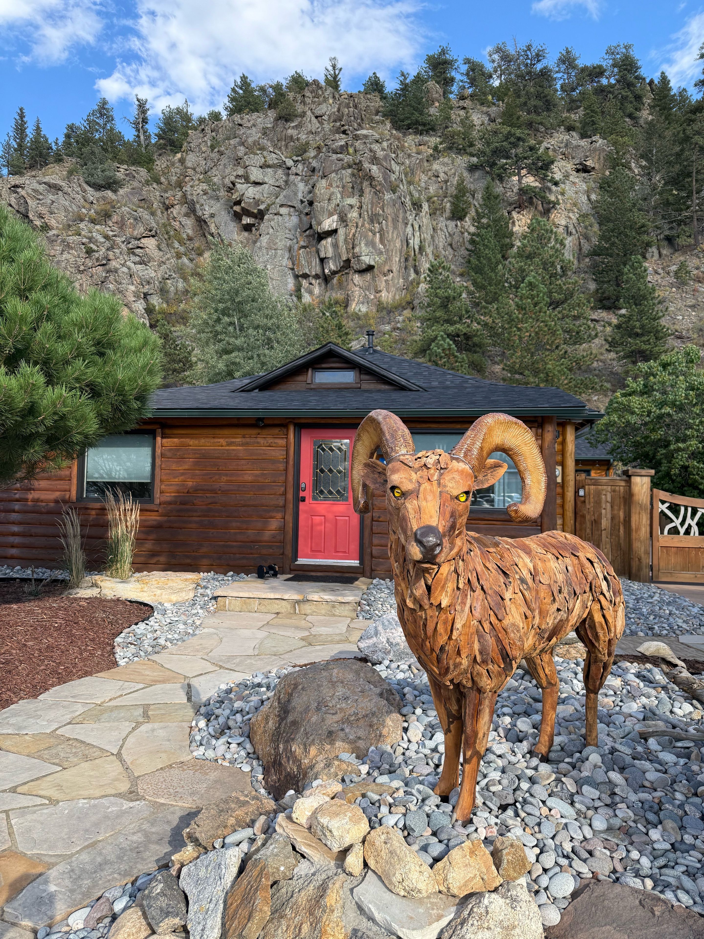 Une cabane en bois avec une porte rouge, une grande sculpture de rocher représentant un bélier, et un paysage montagneux rocheux en arrière-plan.