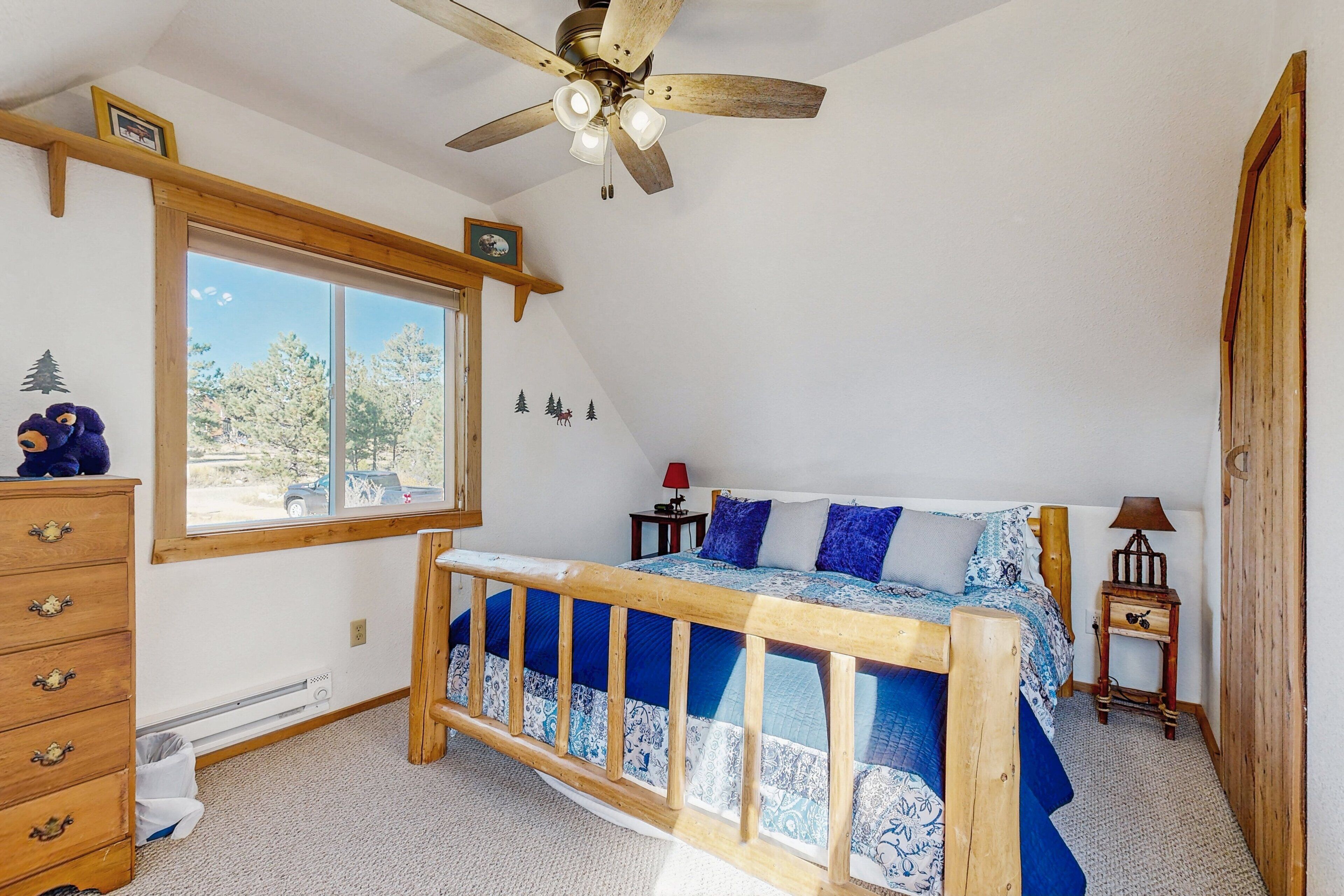 Un dormitorio con cama de madera, ventilador de techo, ventana con vista a árboles, cómoda y mesita de noche con lámpara.