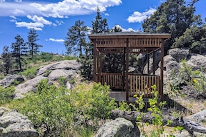 A wooden gazebo with a bench, surrounded by rocks and greenery.