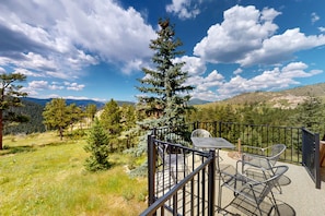 A balcony with a table and chairs, overlooking a mountainous landscape with trees and a clear sky.