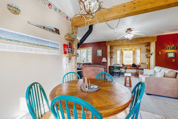 A dining area with a wooden table and turquoise chairs, a ceiling fan, and a red wall with a framed picture. - Spacious Cabin With Stunning Views, Wifi, Fully Stocked Kitchen & W/D (Drake)