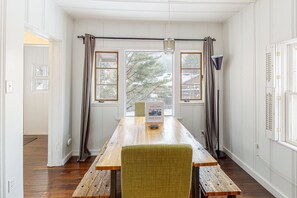 Ein Zimmer mit Holztisch, zwei Stühlen, einem Willkommensschild und Blick auf Bäume durch große Fenster.