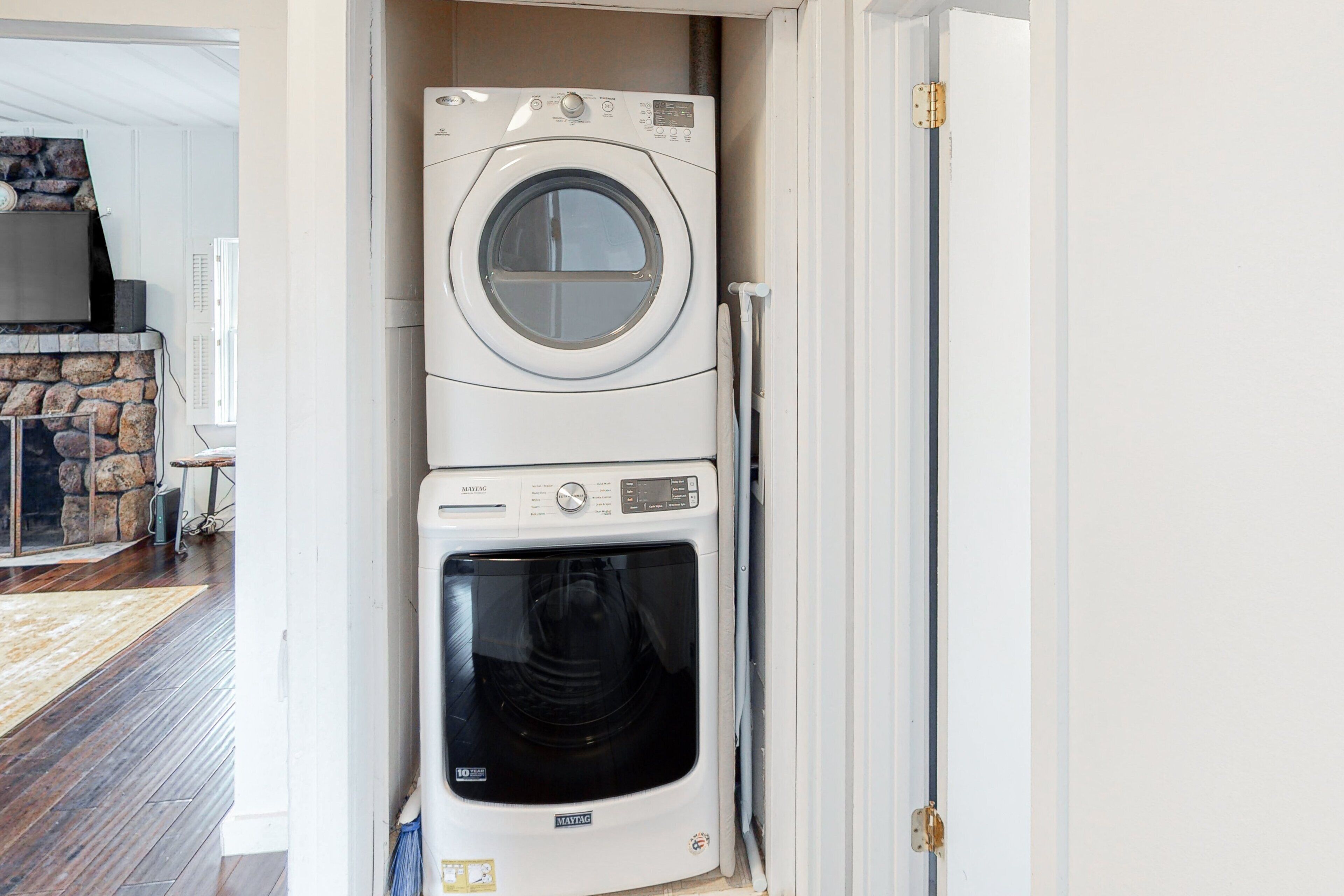 A compact washer and dryer unit in a narrow space, with a stone fireplace and a wooden floor visible in the background.