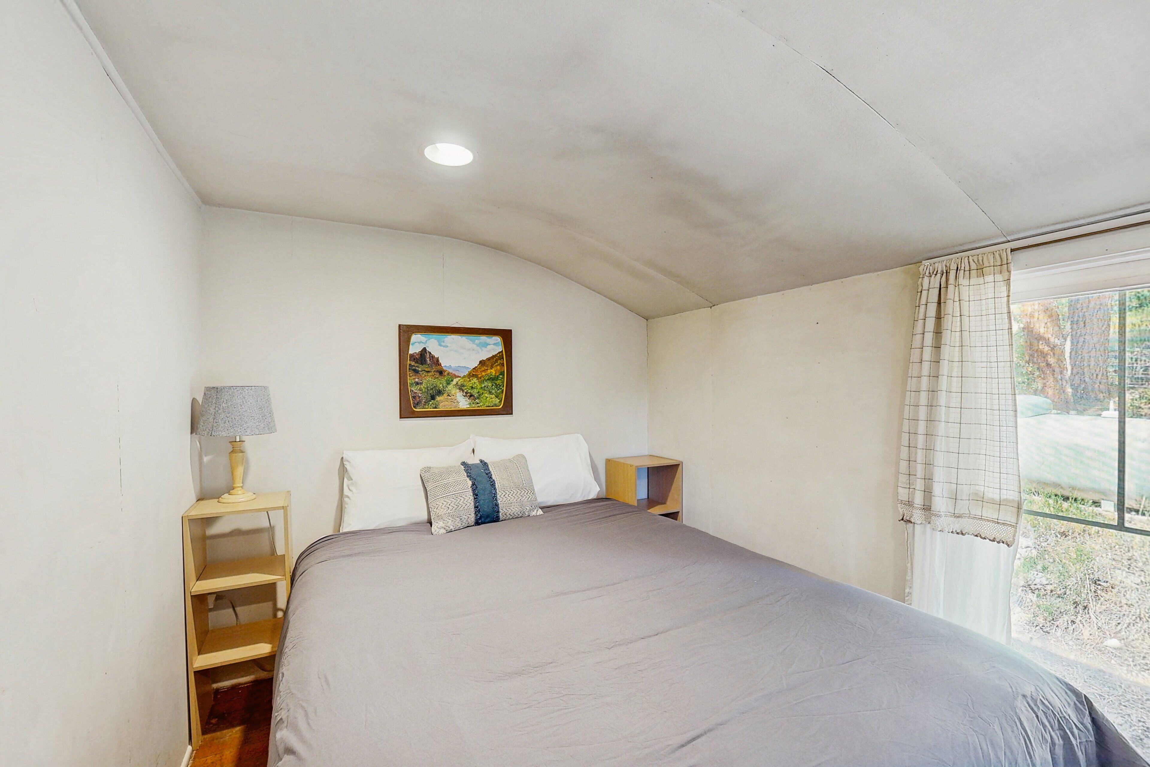 Quarto com cama, mesa de cabeceira, abajur, um quadro na parede e janela com cortinas.